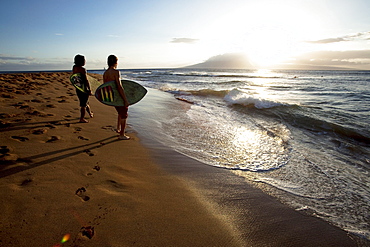 Two men watching the surf at sunset with their skimboards in their arms.