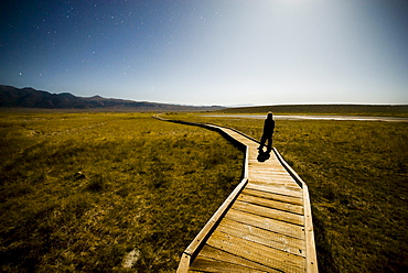 A young man stands on a long wooden pathway that leads to a hot spring.