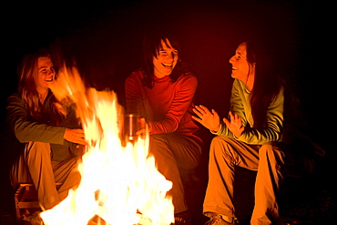 Three women talk and laugh together around a campfire.