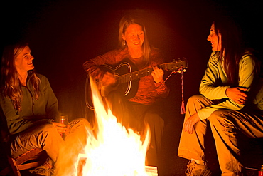 Two women listen, as a third plays the guitar beside a campfire.