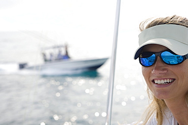 A mid adult woman on a boat is looking at the camera while a fishing boat crosses in the background.