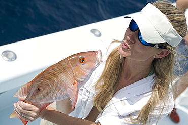 A blonde woman with a white hat and sunglasses holds a freshly caught red snapper and blows it a kiss.