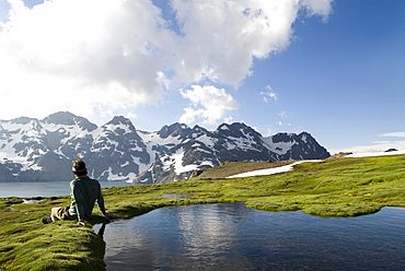 A young man sits next to a small pool enjoying a view of distant snow covered mountains.