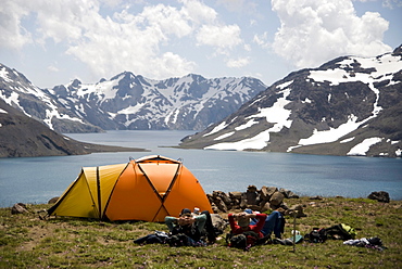 Two men rest next to their orange tent enjoying the dramatic views over a high alpine lake.