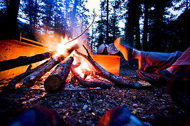 A camper dries her foot next to the flames of their campfire.