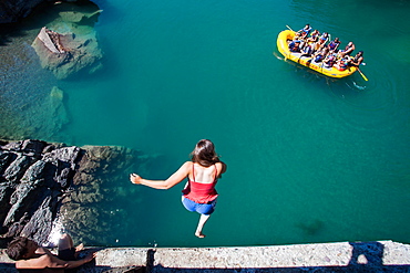 A girl jumps from a high bridge into a river of clear, blue-green water as a raft of people watches from below.