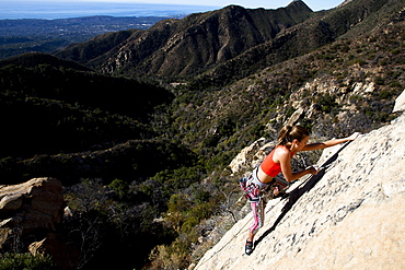 A woman wearing a red tank top and striped pants clips a quick draw while climbing The Rapture (5.8) on Lower Gibraltar Rock in Santa Barbara, California.  The Rapture is a very nice and unbelievably well protected route on the left arête of Lower Gibraltar Rock.