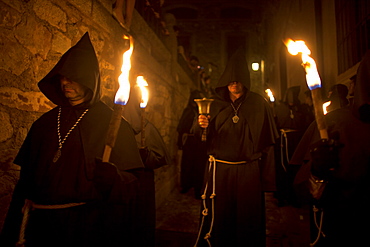 A penitent dressed as a Benedictine monk rings a bell surrounded by men carrying torches at the procession of the Black Christ of Santa Maria during Easter Holy Week inside of the Cathedral of Caceres, Extremadura, Spain