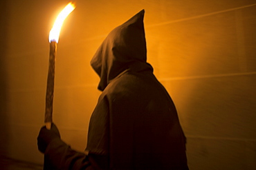 A penitent dressed as a Benedictine monk holds a torch at the procession of the Black Christ of Santa Maria during Easter Holy Week inside of the Cathedral of Caceres, Extremadura, Spain