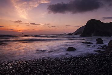 Rocky beach and blurred waves at sunset with Tillamook Rock Lighthouse in distance in Ecola State Park, Oregon.