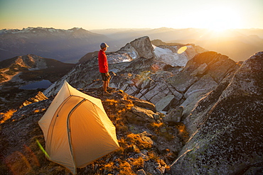 A camper watches the sunset while standing beside his tent on the summit of Saxifrage Peak, Pemberton, BC, Canada.