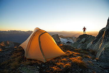 A silhouetted hiker stands on a rock bluff while camping on the summit of Saxifrage Peak, Pemberton, BC, Canada.