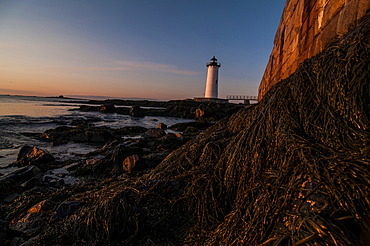 Portsmouth Harbor lighthouse at sunrise with a wall of Fort Constitution lit by the sun.