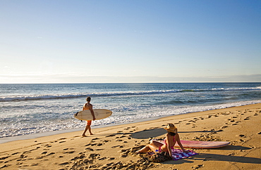 A man carries a surfboard while a young woman in a bikini enjoys the sun setting over the ocean, Mexico