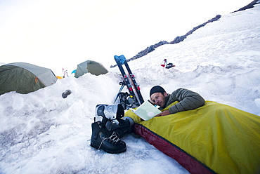 A young climber lies in his bivy sack and reads a book while the sun sets at base camp on Mount Rainier, United States of America