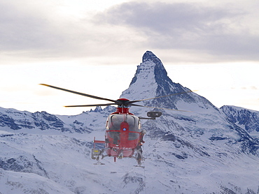 A rescue helicopter of type EC135  is approaching the scene of an accident in the mountains of Zermatt in the Swiss Alps. The famous Matterhorn mountain is in the background.