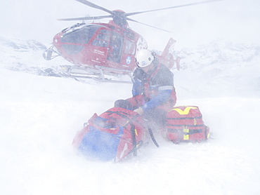 Rescue Personnel is helping an injured skier in the ski area of Zermatt in the Swiss Alps. The emergency doctor protects himself and the patient against the downwash of the landing helicopter.