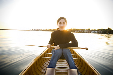 Mature Woman Holds Paddle On Lap In a Wood Canvas Canoe on a calm lake While Looking At Camera With Sun Setting In Background