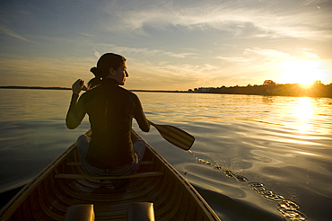 Maure Woman Sits In Front of Wood Canvas Canoe On Lake Looking Off Into The Sunset Holding Paddle