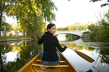 Mature Woman Sitting In a Wood Canvas Canoe in a City Environment Looking Over Shoulder In Between Paddle Strokes