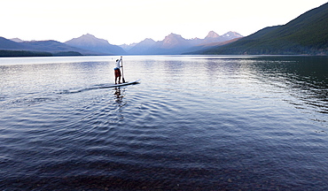 A man stand up paddle boards (SUP) on a calm Lake McDonald in Glacier National Park.