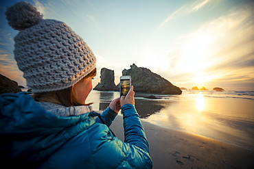 A young woman uses her smartphone to take a picture of the sunset from Bandon Beach, Oregon, United States of America