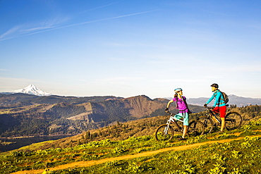 Two young women stop to rest on mountain bikes while riding a single-track trail through an open meadow with river and volcano in distance.