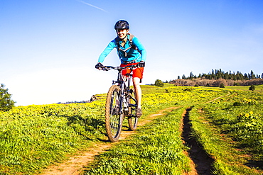 A young woman facing camera rides a mountain bikes down a single-track trail through a meadow.