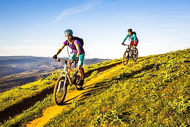 Two young women ride mountain bikes downhill on a single-track trail through green grass in early morning sunlight.