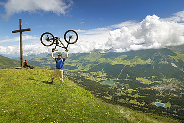 A Mountain Biker Standing On Summit Of Mountain Lifting Mountain Bike Above His Head