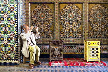 A Distinguished Looking Man Cleans His Glasses While Sitting In Front Of A Spectacularly Patterned Mosaic Wall