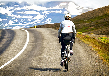 A Cyclist Rides Over The Top Of A Climb With Snowy Mountains In The Background