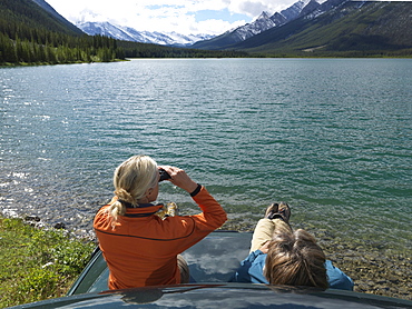 Couple sits on hood of truck and uses binos across lake