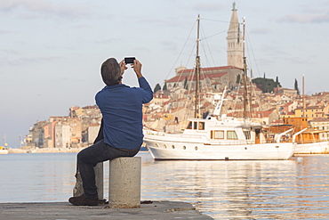 Man takes photo across harbour to old town of Rovinj at sunset