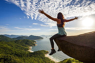 Woman sitting on the edge of the mountain with arms open in Pedra do Telégrafo, Barra de Guaratiba, west side of Rio de Janeiro, Brazil