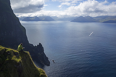 Male hiker sitting on a ledge above the ocean looking at the distant mountains while a seagull passes by.