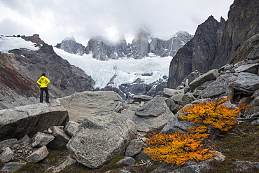 Female hiker with a hand-on-hip pose standing confident on a boulder and looking towards high peaks shrouded in clouds and a glacier below them