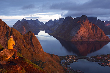 Man with waterproof jacket and beanie sitting on rock looking from a high viewpoint towards mountains and fjords at sunrise. Below, there is a village visible.