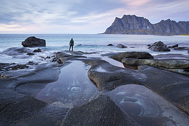 Man with beanie standing at the edge of a rocky beach and looking contemplatively towards a mountain in the distance.