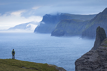 Man standing on the edge of a grassy ledge, with a backpack on his back, looking contemplatively in the distance.