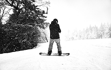 Black and white image of a snowboarder stopped on the edge of a trail.