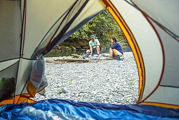 Two hikers tend to a campfire next to their tent on a riverbed.