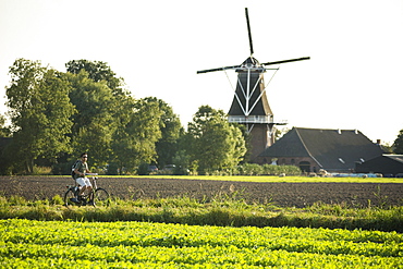 A young man wearing shorts, a green t-shirt and sunglasses, rides his bike on a sunny day, in the Dutch countryside, with a windmill in the background, in the province of Groningen, in The Netherlands.