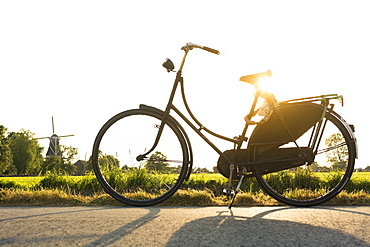 A vintage, worn traditionally Dutch bicycle propped up on the side of a bikepath, in the Dutch countryside at sunset, with a windmill in the background, in the province of Groningen, The Netherlands.