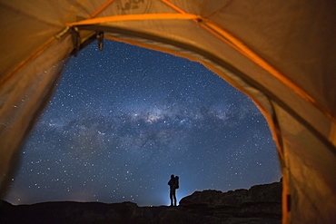 Silhouette of backpacker standing against starry night sky and Milky Way galaxy visible from inside of pitched tent, New South Wales, Australia