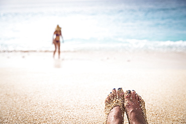 Feet of woman covered with sand on beach and woman in bikini in background