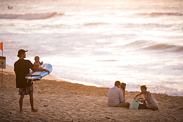 Surfer with baby on surfboard and other people on beach at sunset, Oahu, Hawaii Islands, USA