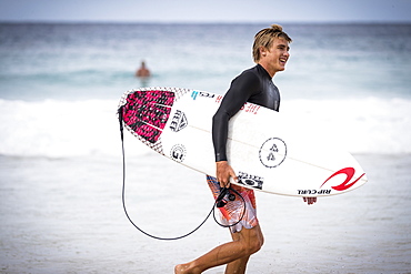 Side view of single smiling male surfer carrying surfboard along beach