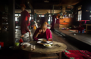 Young monk eating at table in rural monastery with another monk walking behind in background, Myanmar, Shan, Myanmar