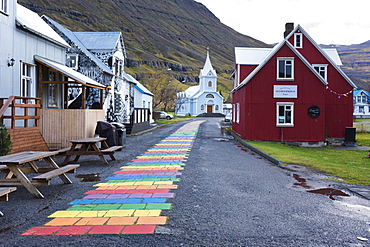 Rainbow colored brick road between houses leading to small church at edge of town, Seydisfjordur, Iceland